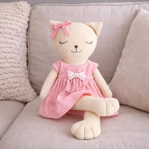Könnte beinhalten: Eine Plüschkatzenpuppe mit geschlossenen Augen, die ein rosa Kleid mit einer weißen Schleife trägt. Die Puppe hat eine rosa Schleife auf dem Kopf und sitzt auf einem grauen Sofa. Die Pfoten der Katze sind gekreuzt.