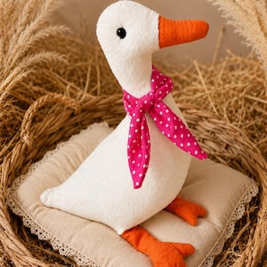 May include: A plush white goose toy with an orange beak and orange webbed feet. The goose wears a bright pink neckerchief featuring a white polka dot pattern. It has a small black bead eye and is positioned sitting on a square beige fabric cushion.