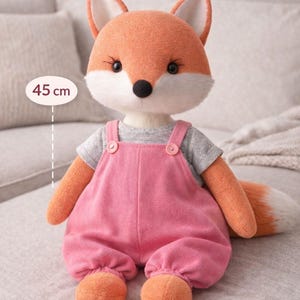Puede incluir: Un peluche de zorro, de aproximadamente 45 cm de altura, con un mono rosa y una camiseta gris. El zorro tiene pelaje naranja, detalles blancos y una nariz negra. La imagen incluye el texto "PDF Pattern + Video Tutorial" y "Fox Plush Pattern".