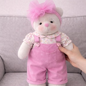 May include: A plush white teddy bear wearing a pink bow, headband, and overalls. The bear has rosy cheeks, a pink nose, and a floral print blouse. The overalls have heart-shaped accents.
