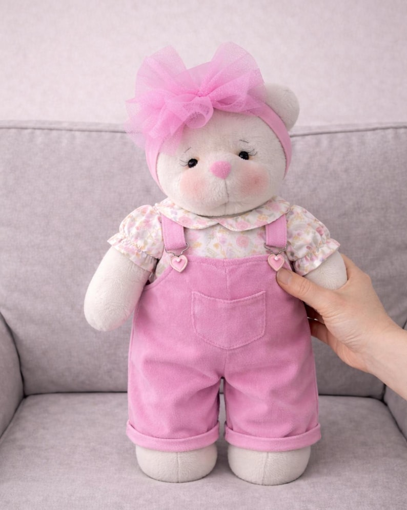 May include: A plush white teddy bear wearing a pink bow, headband, and overalls. The bear has rosy cheeks, a pink nose, and a floral print blouse. The overalls have heart-shaped accents.