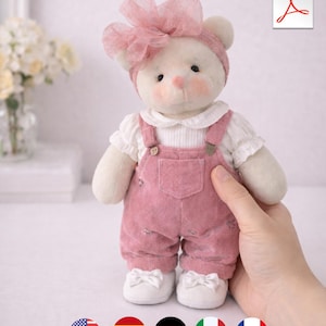 May include: A handmade plush bear wearing a pink corduroy overall, white collared shirt, and white shoes with bows. The bear has a pink bow and headband. The bear is being held by a hand.