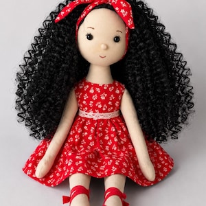May include: A fabric doll with long curly black hair, wearing a red dress with a white floral pattern, a matching red headband, and red shoes. The doll measures 50 cm in height. A PDF icon is visible in the corner of the image.