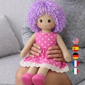 Könnte beinhalten: Eine handgemachte Puppe mit Stoffkörper, die ein rosa gepunktetes Kleid mit Spitzenbesatz trägt. Die Puppe hat lila Garnhaare und rosa Schuhe. Die Puppe ist 45 cm groß.