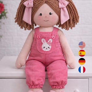 Könnte beinhalten: Eine handgefertigte Puppe, 55 cm groß, mit braunem Garnhaar, das zu Zöpfen mit rosa Schleifen gestylt ist. Die Puppe trägt eine rosa Latzhose mit einem Hasenaufnäher und passenden Schuhen mit Schleife. Die Puppe hat ein lächelndes Gesicht.
