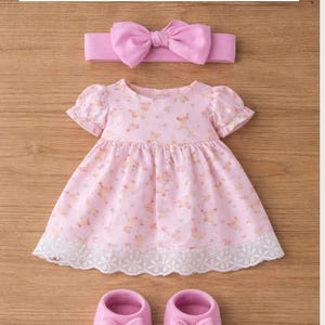 May include: A pink baby outfit set, including a dress, shoes, and headband. The dress features a floral pattern and lace trim. The shoes and headband have bow accents. The text at the top reads "Roupinha 1: Vestido + Sapato + La&ccedil;o".