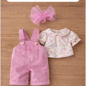 May include: A pink corduroy jumpsuit with heart-shaped clasp details, a floral print blouse, and a pink tulle bow. The outfit is displayed on a wooden surface. Text on the image reads: Outfit 2: Jumpsuit + Blouse + Bow.