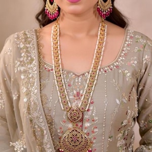 May include: A gold-toned necklace with a large pendant and matching earrings. The necklace features multiple strands of white beads and red gemstones. The earrings and pendant have a detailed design with red accents. The woman is wearing a light green dress with floral embroidery.