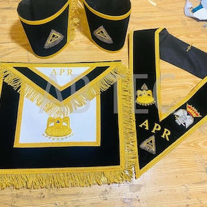 Puede incluir: Conjunto de insignias masónicas en negro y dorado, que incluye un delantal, un collar y puños. El delantal presenta un centro blanco con flecos y bordados dorados, y las letras "APR". El collar y los puños son negros con detalles dorados.