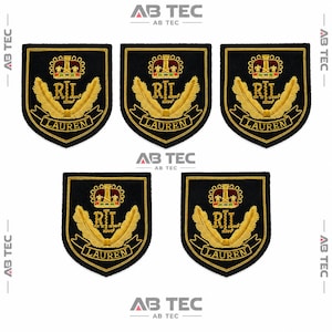Op de afbeelding: Vijf zwarte, schildvormige geborduurde patches met een gouden kroon boven de letters RIL, geflankeerd door twee gouden vleugels, met het woord LAUREN in goud geborduurd op een banner aan de onderkant. Elke patch heeft een zwarte rand.