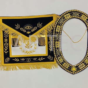 Puede incluir: Un conjunto de delantal y collar masónico negro y dorado. El delantal presenta flecos dorados, bordados y un panel blanco con símbolos. El collar es ovalado, negro con adornos dorados y una cadena dorada.
