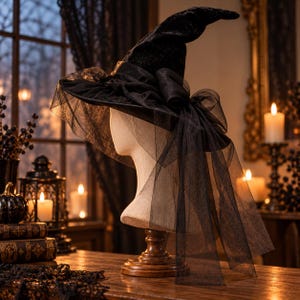 Puede incluir: Un sombrero de bruja negro con una copa puntiaguda y ala ancha, adornado con capas de tul negro transparente y un gran lazo decorativo. El sombrero se exhibe sobre un soporte de cabeza de maniquí beige, mostrando la textura de la tela.