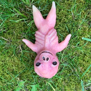 May include: A pink, stylized sculpture of a creature with a humanoid form, lying on a bed of green grass and moss. The sculpture has a detailed face with dark eyes and a smiling expression. The arms are outstretched, and the tips of the ears are white.