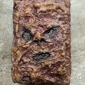 May include: A rectangular, textured prop book with a grotesque, weathered appearance. The cover features a sculpted face with dark eye sockets and a gaping mouth. The color palette includes shades of brown, red, and gold, creating a disturbing, aged effect.