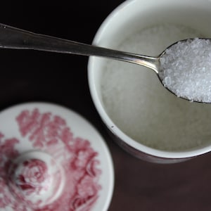 Puede incluir: Un azucarero de cerámica blanca con un patrón floral rojo de transferencia en la tapa y el lateral. Una cuchara de metal plateado descansa sobre la parte superior del cuenco, llena de azúcar granulado blanco, mientras la tapa a juego está cerca.