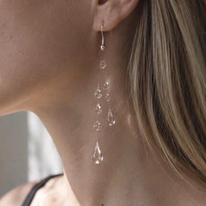 May include: A long drop earring featuring a silver hook and a delicate chain adorned with multiple clear, teardrop-shaped glass beads of varying sizes that hang vertically, creating a cascading effect along the side of the neck.