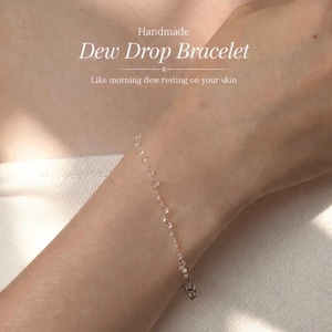 May include: A delicate bracelet featuring small, clear, dew-drop-like beads strung along a thin chain. The image includes the text: Handmade Dew Drop Bracelet, Like morning dew resting on your skin. The bracelet is worn around a wrist.
