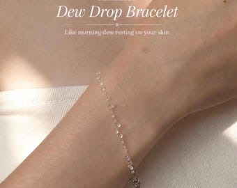Könnte beinhalten: Ein zartes Armband mit kleinen, klaren, tau-tropfenartigen Perlen an einer dünnen Kette. Das Bild enthält den Text: Handmade Dew Drop Bracelet, Like morning dew resting on your skin. Das Armband wird um ein Handgelenk getragen.