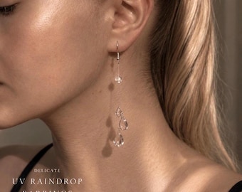 Könnte beinhalten: Hängeohrringe bestehend aus einem dünnen silberfarbenen Draht mit einer kleinen klaren Perle und zwei klaren tropfenförmigen Anhängern. Der Text DELICATE UV RAINDROP EARRINGS steht links. Die Ohrringe hängen an einem einfachen silberfarbenen Haken.