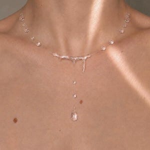 Può includere: Una collana trasparente caratterizzata da un elemento centrale a forma di ramo con un filo verticale pendente che termina a goccia. La catena è composta da un cordino sottile e quasi invisibile, arricchito da piccole perle sferiche.