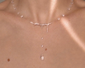Könnte beinhalten: Eine klare, transparente Halskette mit einer Reihe kleiner, tropfenförmiger Perlen, die auf einer dünnen, unsichtbaren Schnur aufgereiht sind. In der Mitte befindet sich ein größeres, unregelmäßiges, geschmolzen wirkendes Anhängerelement.