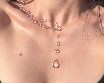 Könnte beinhalten: Eine transparente Halskette mit einer Reihe kleiner, tropfenförmiger Perlen, die auf einer dünnen, unsichtbaren Schnur aufgereiht sind. Das Design umfasst eine mittlere Schlaufe und einen vertikalen Anhänger, der in einer größeren Tränenform endet.