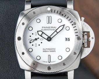 May include: A stainless steel Panerai Submersible automatic watch with a white dial, black hands, and a date display. The watch has a black rubber strap and is water-resistant to 300m/1000ft. The watch face includes the text "PANERAI SUBMERSIBLE AUTOMATIC 300m/1000ft".