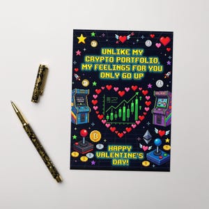 May include: A Valentine's Day card with a crypto theme. The card features pixelated graphics of arcade games, hearts, and cryptocurrency symbols. Text on the card reads, "Unlike my crypto portfolio, my feelings for you only go up!" A gold and black pen and a matching pen are next to the card.