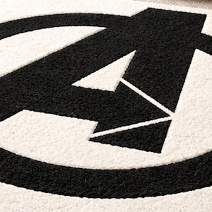 Könnte beinhalten: Ein schwarz-weißer Teppich mit dem Avengers-Logo. Das Logo ist ein stilisiertes "A" in einem Kreis. Der Teppich hat ein strukturiertes, weiches Aussehen, mit dem Design in Schwarz auf weißem Hintergrund. Dieser Teppich ist für ein Wohn- oder Schlafzimmer geeignet.