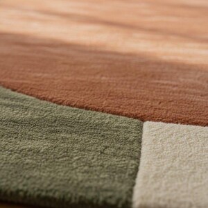 May include: Close-up of a textured area rug with a geometric design. The rug features sections of muted colors, including shades of brown, green, and off-white. The soft, plush texture is visible, suggesting a comfortable and inviting feel.