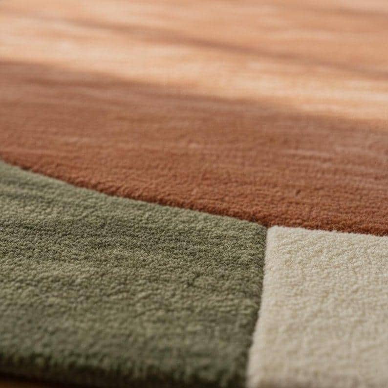 May include: Close-up of a textured area rug with a geometric design. The rug features sections of muted colors, including shades of brown, green, and off-white. The soft, plush texture is visible, suggesting a comfortable and inviting feel.