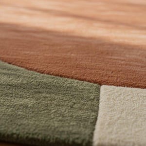 May include: Close-up of a textured area rug with a geometric design. The rug features sections of muted colors, including shades of brown, green, and off-white. The soft, plush texture is visible, suggesting a comfortable and inviting feel.