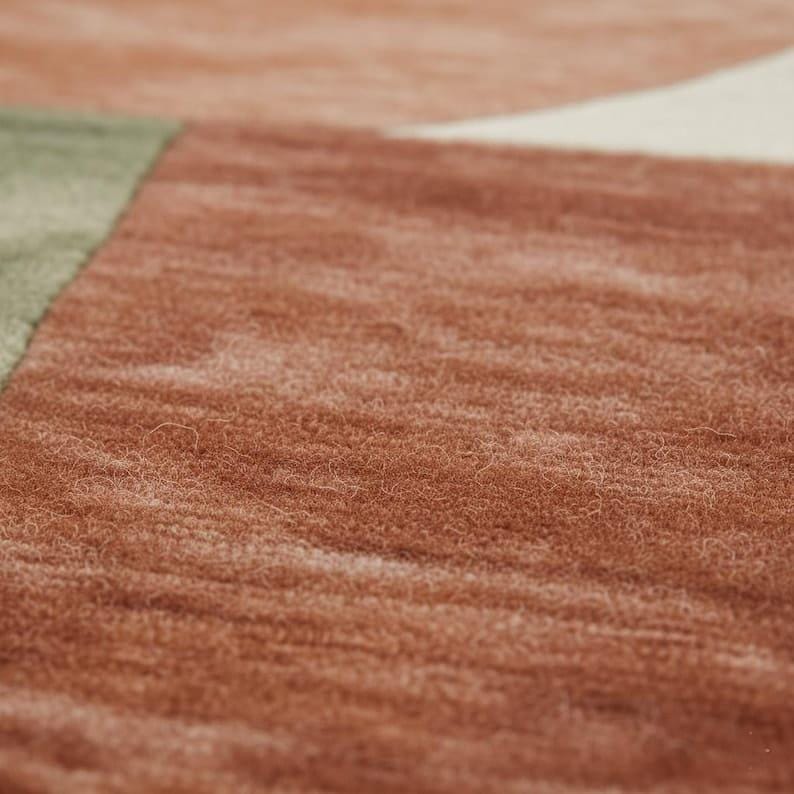 May include: Close-up of a textured rug with a geometric design. The rug features a color palette of rust, cream, and sage green. The fibers have a soft, plush appearance, suggesting a comfortable and inviting texture.