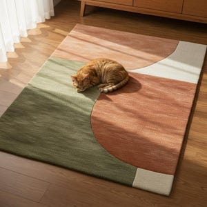 May include: A rectangular area rug with a geometric design in shades of green, peach, and white. An orange tabby cat is curled up on the rug. The rug is placed on a hardwood floor near a wooden cabinet.