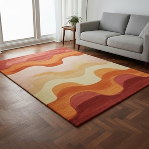 Könnte beinhalten: Rechteckiger Teppich mit wellenförmigem Muster in Rot-, Orange- und Cremetönen. Der Teppich liegt auf einem Holzboden und hat eine weiche, strukturierte Oberfläche. Das Design erinnert an einen Sonnenuntergang oder eine Wüstenlandschaft.