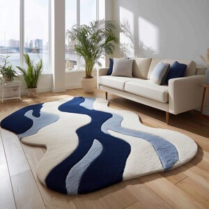 Könnte beinhalten: Abstrakter Teppich mit fließendem Design in Weiß, Hellblau und Marineblau. Der Teppich liegt auf einem Holzboden in einem Wohnzimmer, mit einem weißen Sofa und großen Fenstern im Hintergrund. Das Design des Teppichs ähnelt einem Fluss.