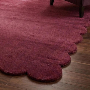 May include: A deep burgundy rug with a scalloped edge, placed on dark wood flooring. The rug's plush texture is evident. The edge of a purple armchair is visible, suggesting a warm and inviting interior setting.