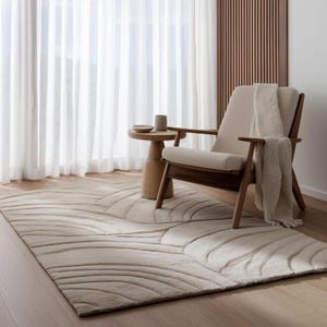May include: A neutral-toned area rug with a wave-like pattern dominates the foreground. A wooden chair with a cream-colored cushion and a matching throw blanket sits to the right. A small wooden side table holds a cup and a dish. Sheer white curtains and a wooden wall panel are in the background.