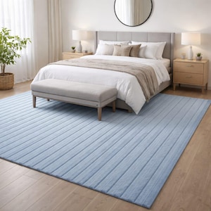 Peut inclure: Un tapis rectangulaire bleu clair avec un motif côtelé est posé sur un parquet dans une chambre. Le tapis est devant un lit avec une literie blanche, une tête de lit grise et un banc. Deux tables de chevet avec des lampes encadrent le lit.