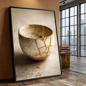 Może przedstawiać: Oprawiony wydruk ceramicznej miski, prezentujący japońską sztukę Kintsugi. Miska jest w kolorze złamanej bieli z pęknięciami wypełnionymi złotem. Dzieło sztuki jest osadzone na neutralnym tle, z widokiem na miasto widocznym przez okno.