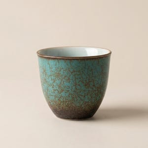 May include: A teal ceramic cup with a speckled texture and a dark brown base. The interior is a light blue color, and the rim is a dark brown. The cup has a smooth, rounded shape.