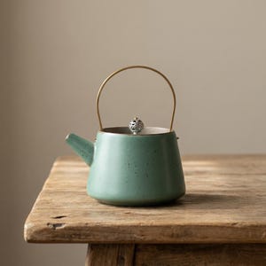 Puede incluir: Una pequeña tetera de cerámica color turquesa con asa dorada y tapa plateada decorativa. La tetera está moteada y se encuentra sobre una mesa de madera rústica, mostrando un diseño minimalista.