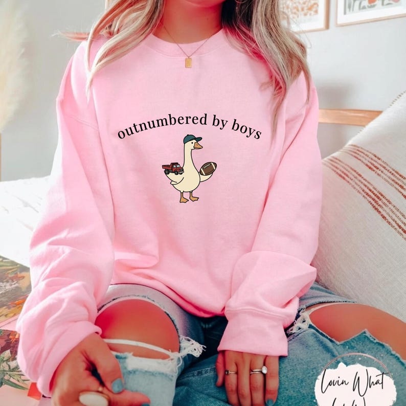 May include: Pink sweatshirt with the phrase "outnumbered by boys" in black lettering. A cartoon goose wearing a baseball cap and holding a football and a toy truck is printed on the front. The sweatshirt is a crew neck style.