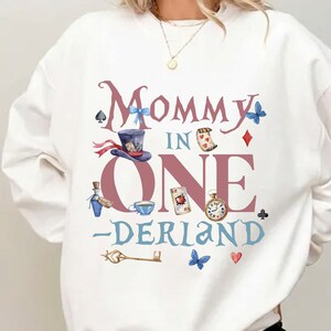May include: White sweatshirt with a whimsical "Mommy in Onerland" design. Features illustrations of a hat, playing cards, a pocket watch, and other Alice in Wonderland-themed elements. The design is in shades of pink, blue, and brown.