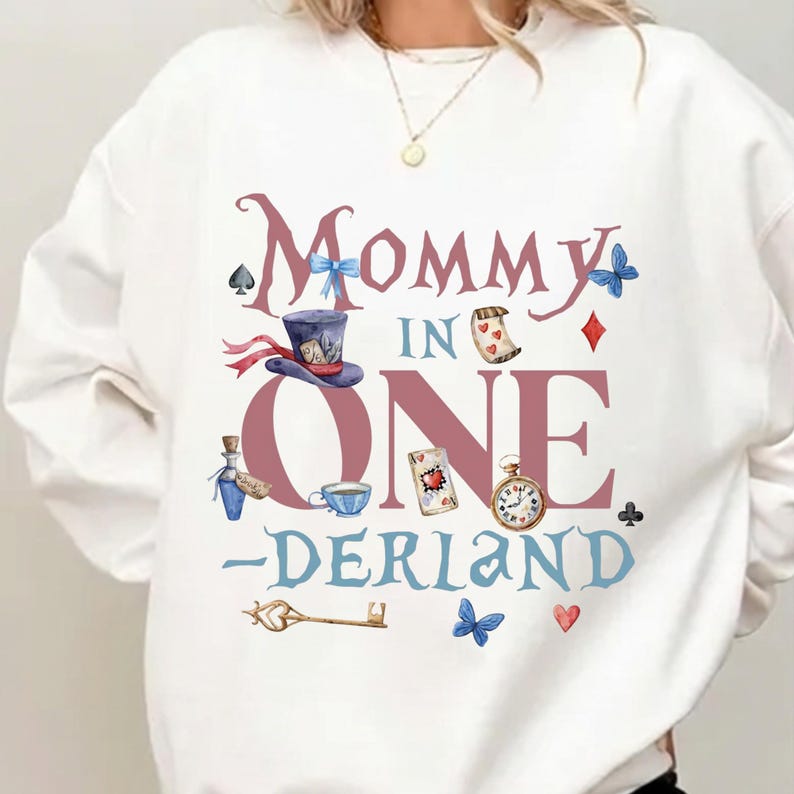 May include: White sweatshirt with a whimsical "Mommy in Onerland" design. Features illustrations of a hat, playing cards, a pocket watch, and other Alice in Wonderland-themed elements. The design is in shades of pink, blue, and brown.