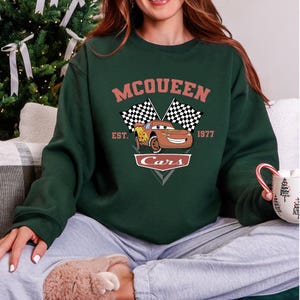 May include: Dark green sweatshirt featuring a graphic of Lightning McQueen from the movie Cars. The design includes the text "McQueen" above the car, with checkered flags and the text "Est. 1977 Cars" below. The sweatshirt is a crew neck style.