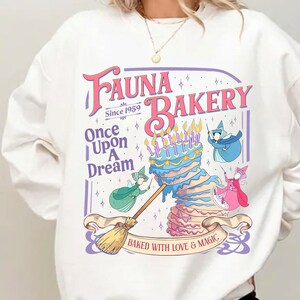 May include: A white crewneck sweatshirt featuring a graphic print that reads Fauna Bakery Since 1959, Once Upon A Dream, and Baked With Love & Magic. The design includes a multi-layered cake with candles and three fairy figures in colorful robes.