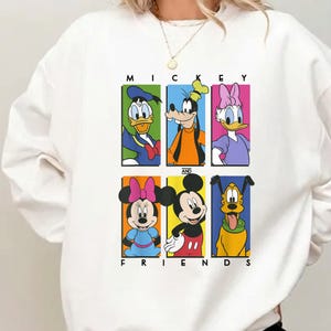 May include: White sweatshirt featuring a colorful graphic of Mickey Mouse and friends. The design includes Donald Duck, Goofy, Daisy Duck, Minnie Mouse, Mickey Mouse, and Pluto, with the words "MICKEY AND FRIENDS" printed above and below the characters.
