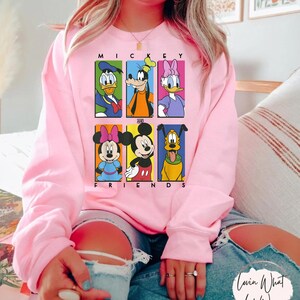 May include: A light pink sweatshirt featuring a graphic of Mickey Mouse and friends. The design includes Donald Duck, Goofy, Daisy Duck, Minnie Mouse, Mickey Mouse, and Pluto. The words "MICKEY AND FRIENDS" are printed above and below the characters.