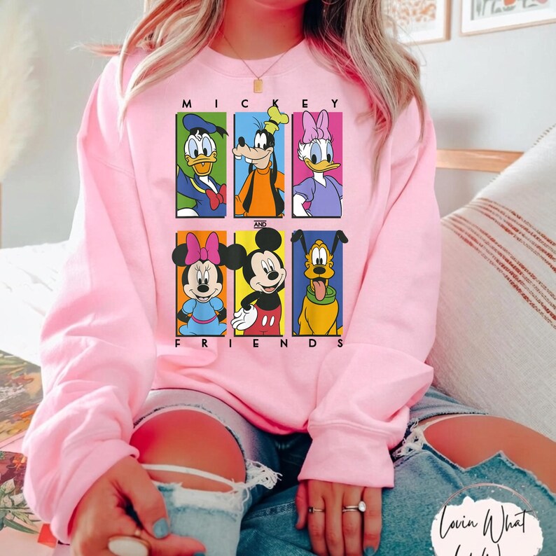 May include: A light pink sweatshirt featuring a graphic of Mickey Mouse and friends. The design includes Donald Duck, Goofy, Daisy Duck, Minnie Mouse, Mickey Mouse, and Pluto. The words "MICKEY AND FRIENDS" are printed above and below the characters.